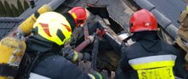 Prace rozbiórkowe nadpalonego dachu budynku mieszkalnego. Czterech strażaków na dachu prowadzi akcję gaśniczą.