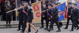 poczet sztandarowy składający się z kilkunastu strażaków ubranych w umundurowanie wyjściowe z czapka rogatywką na głowie maszeruje przed trybuną honorową, goście w tle stoją na baczność, strażacy salutują do sztandaru