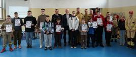 Na zdjęciu znajduje się grupa młodzieży i dorosłych, pozująca wspólnie do zdjęcia. Uczestnicy trzymają w rękach dyplomy i nagrody, . W tle widać umundurowanych strażaków. 