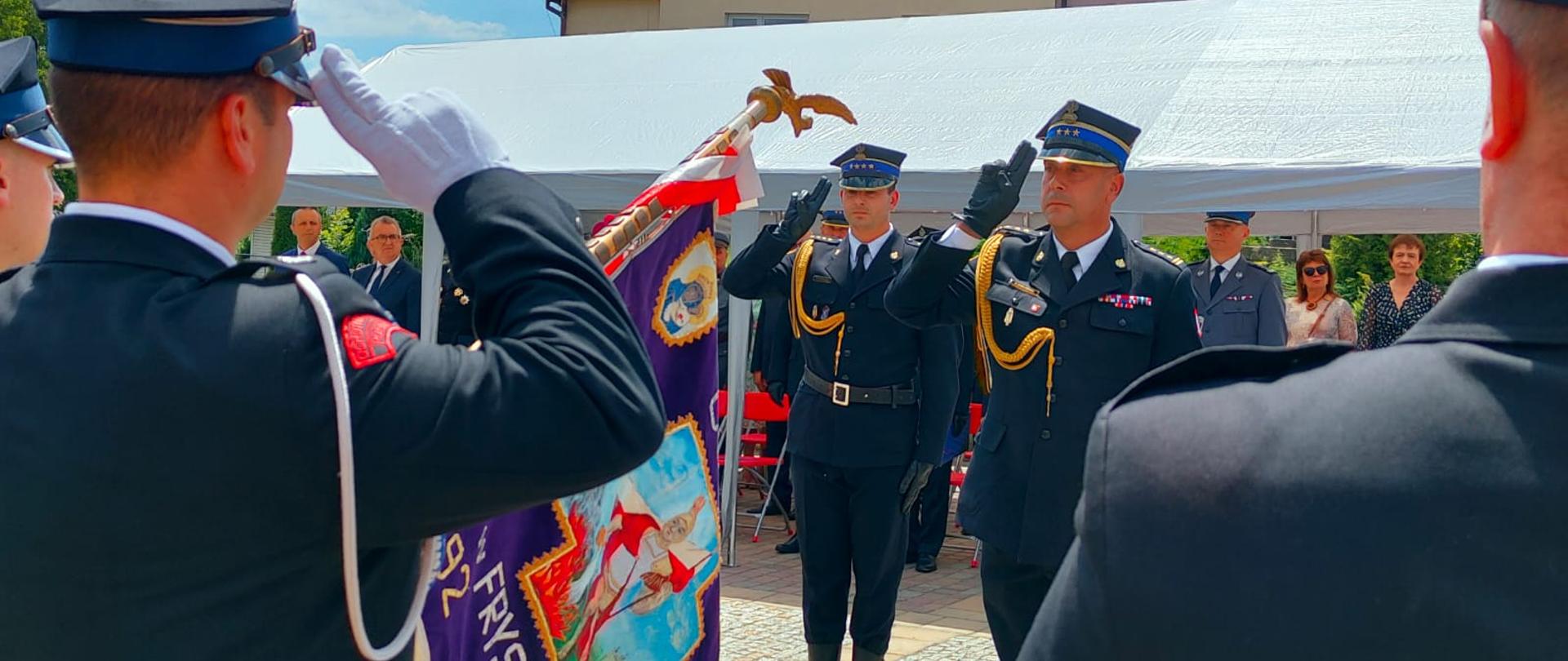 Zdjęcie zrobione na zewnątrz podczas uroczystego apelu. Dwóch oficerów Państwowej Straży Pożarnej w mundurach galowych salutuje pocztowi sztandarowemu.