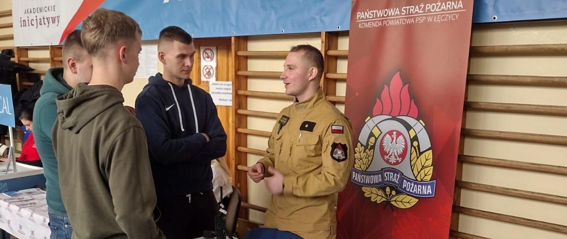Na zdjęciu widać strażaka w piaskowym stroju, który trzem uczniom coś opowiada. Przed nimi stoi stolik, na którym znajduje się defibrylator i worek AMBU. Na podłodze leży fantom do ćwiczeń. Za strażakiem jest baner z logo Państwowej Straży Pożarnej i napisem Państwowa Straż Pożarna Komenda Powiatowa Państwowej Straży Pożarnej w Łęczycy. Za banerem na drabinkach do ćwiczeń, na której powieszony jest baner z napisem Akademickie.