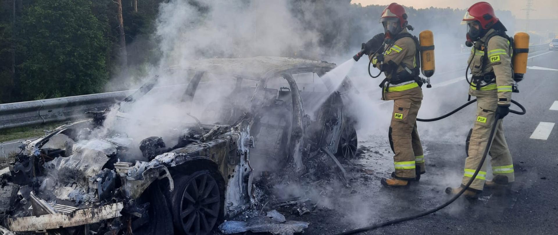 Na zdjęciu widać doszczętnie spalony samochód osobowy na trasie S7. Obok niego stoją strażacy ubrani w ubrania bojowe i aparaty ochrony układu oddechowego dogaszający pożar. W tle widać drzewa i infrastrukturę drogową.