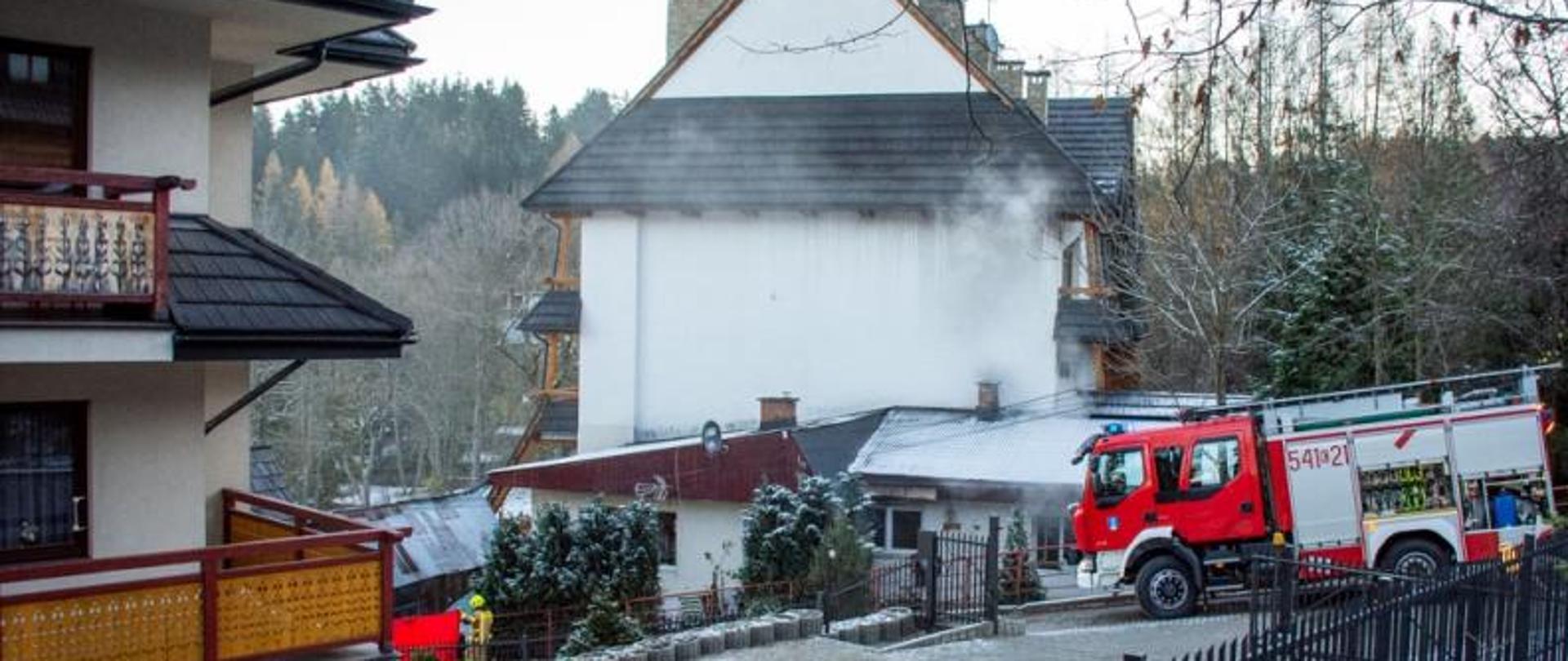 Zdjęcie przedstawia pożar budynku mieszkalnego w Zakopanem na ul. Jaszczurówka. Z budynku wydobywa się dym, przed budynkiem stoi wóz strażacki. W tle widać las.