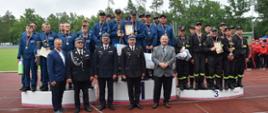 Trzy drużyny męskie stoją na podium. Wszyscy mają medale zawieszone na szyj. Przed podium stoi pięciu mężczyzn. Trzech w środku to strażacy w mundurach wyjściowych. W tle widać zieloną trawę i drzewa.