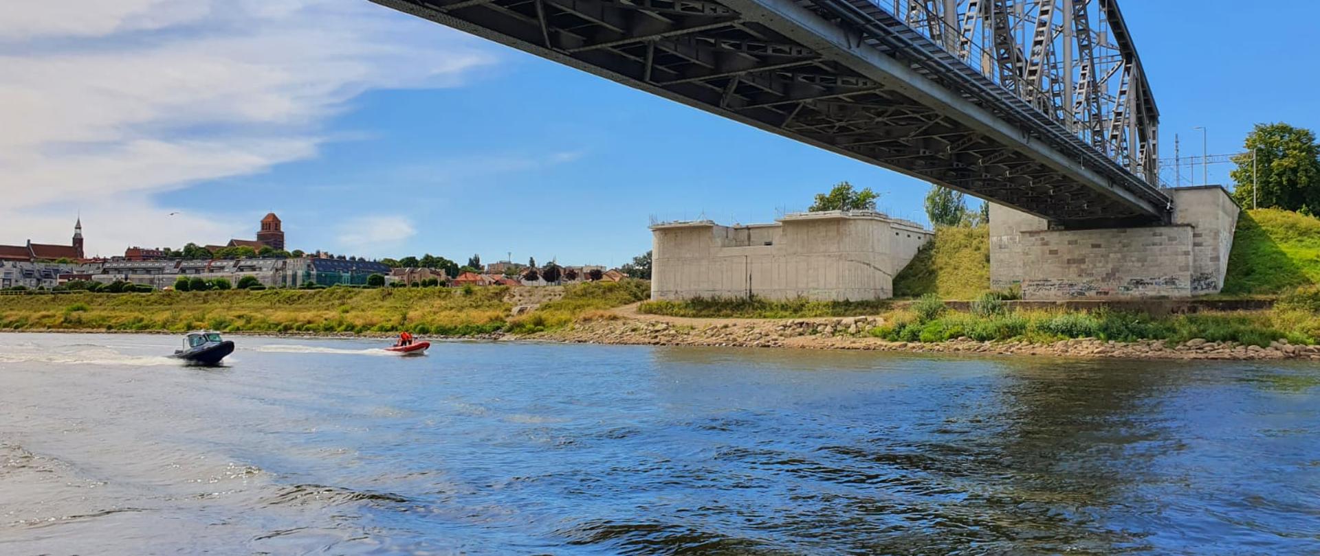 Widok z brzegu Wisły. Na wodzie dwie motorówki - strażacka i policyjna przepływają pod kolejowym mostem lisewskim - konstrukcja stalowej kratownicy, po drugiej stronie brzegu widać starówkę Tczewa.