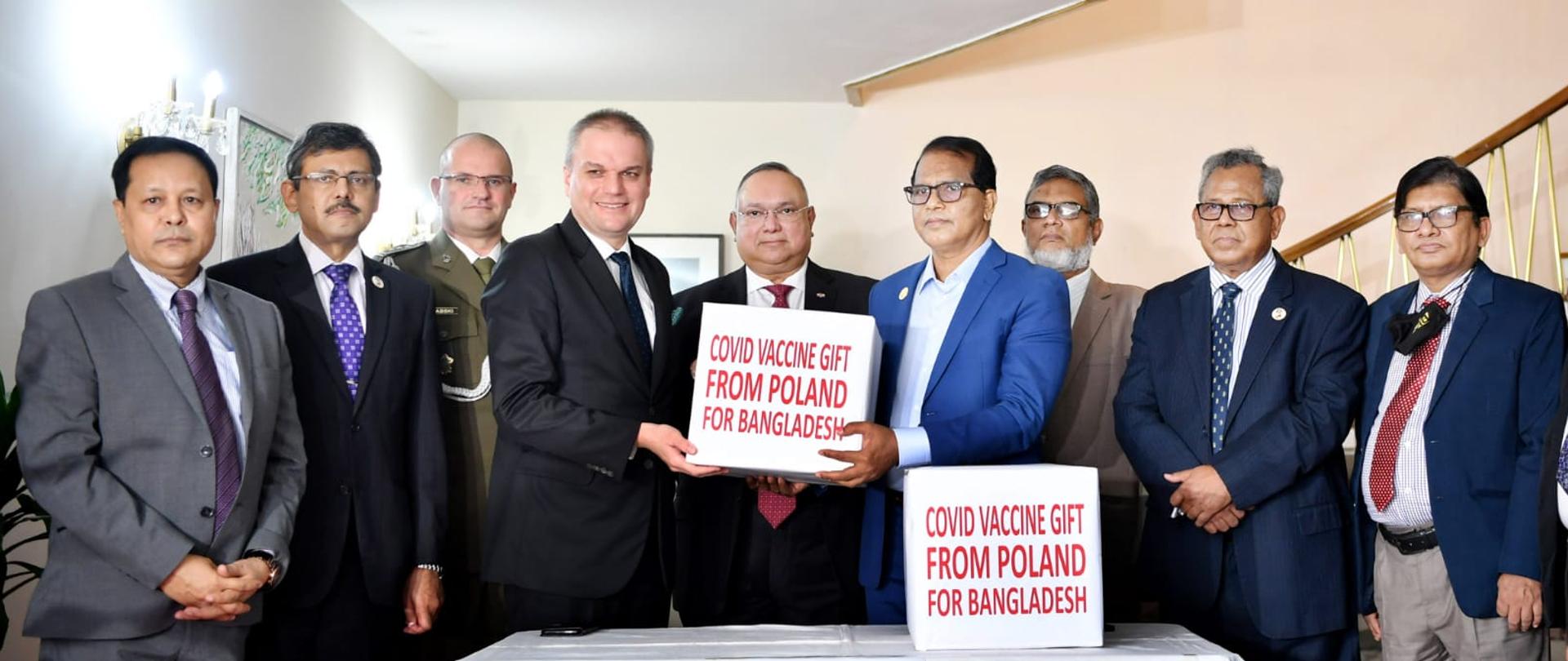 Grupa mężczyzn stoi za stołem. Jeden mężczyzna przekazuje drugiemu mężczyźnie pudełko z napisem "Covid vaccine gift from Poland for Bangladesh"
