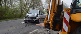 Zderzenie auta z koparko-ładowarką. Droga krajowa nr 36 pomiędzy Wodnikami i Załęczem. Na pierwszym planie - fragment koparko-ładowarki oraz auto koloru srebrnego uczestniczące w wypadku. Pojazd ma bardzo mocno uszkodzoną przednią część. Wokół samochodu kilku strażaków. Na jezdni porozrzucane są fragmenty karoserii auta. W tle drzewa.