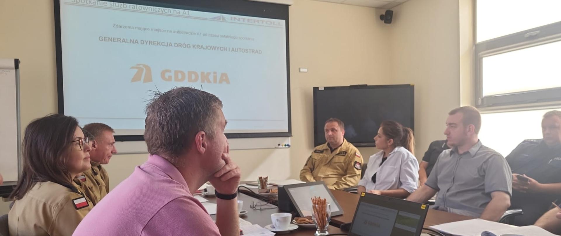 Spotkanie służb w sprawie bezpieczeństwa ruchu drogowego na autostradzie A1. 9 osób na zdjęciu, w tym policjanci, strażacy i osoby cywilne podczas spotkania. W tle ekran z prezentacją GDDKiA.
