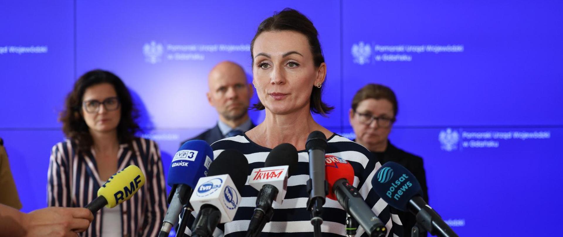 Kobieta stoi przed mikrofonami , w tle inne osoby. Na tablicy napis Pomorski Urząd Wojewódzki w Gdańsku