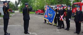 Widok z boku. Małopolski komendant wojewódzki PSP stoi przed pocztem sztandarowym i salutuje. Za nim salutuje strażak - dowódca uroczystości. Poczet sztandarowy pochyla sztandar