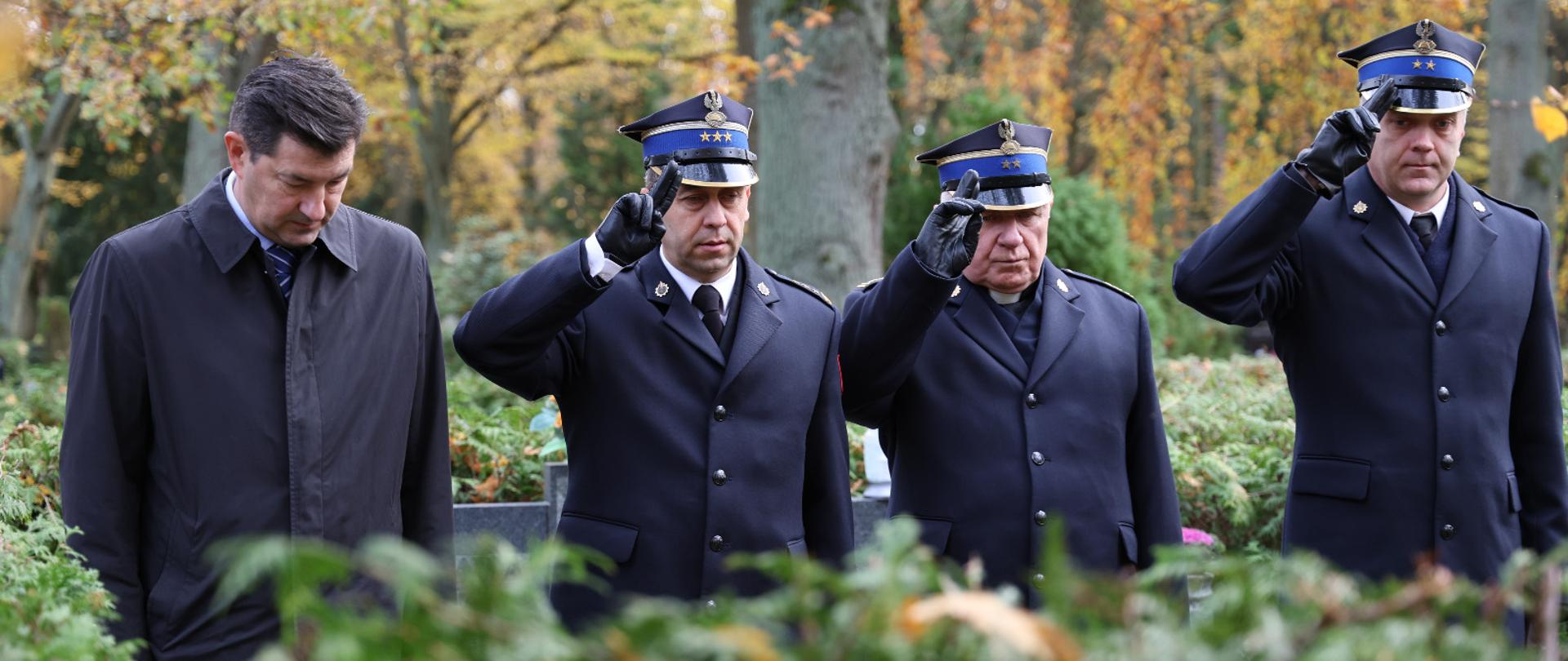 Czterech mężczyzn stoi na cmentarzu wśród jesiennych drzew. Trzech z nich w mundurach strażackich salutuje, czwarty ubrany w płaszcz stoi obok. Tło rozmyte, widać krzewy i nagrobki.