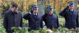 Czterech mężczyzn stoi na cmentarzu wśród jesiennych drzew. Trzech z nich w mundurach strażackich salutuje, czwarty ubrany w płaszcz stoi obok. Tło rozmyte, widać krzewy i nagrobki.