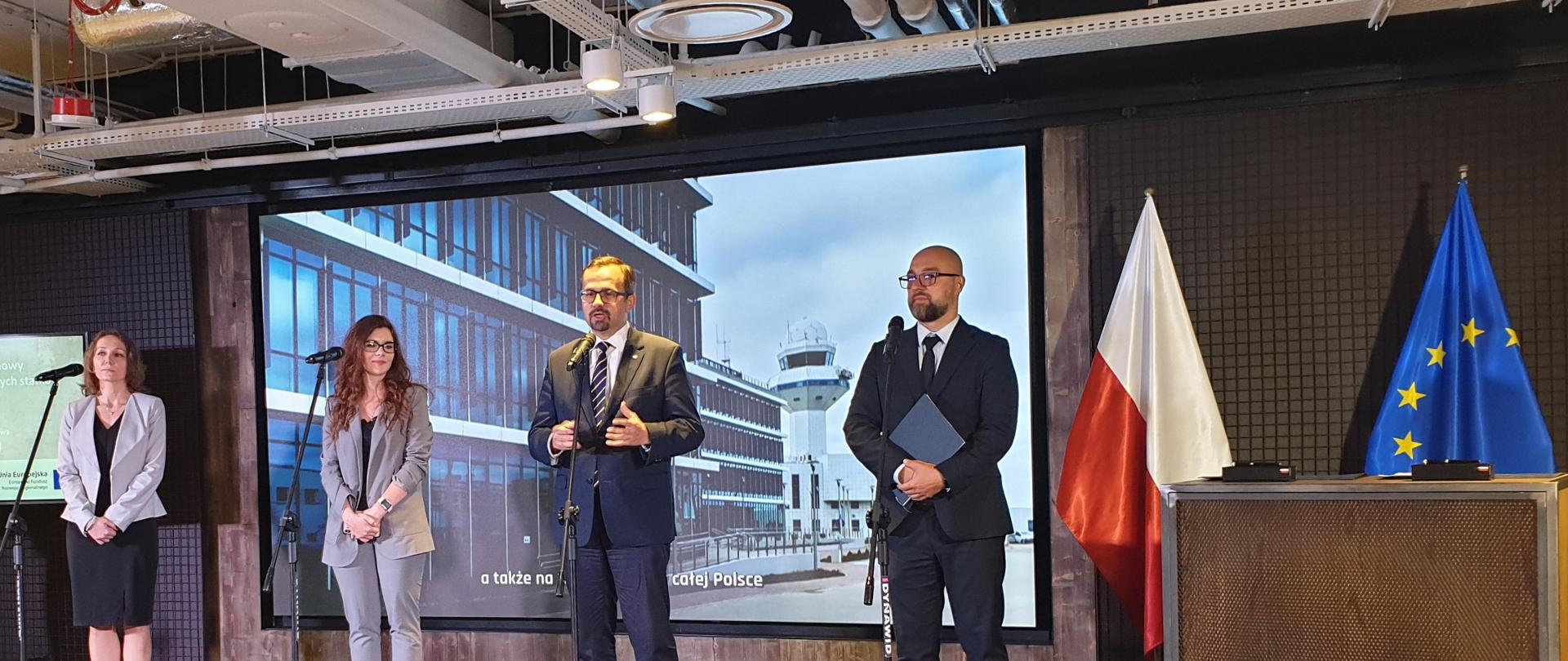 Na zdjęciu widać 4 osoby, które stoją na podwyższeniu. Wszyscy stoją przy mikrofonach. W ich tle, po prawej stronie stoją flagi Polski i Unii Europejskiej.