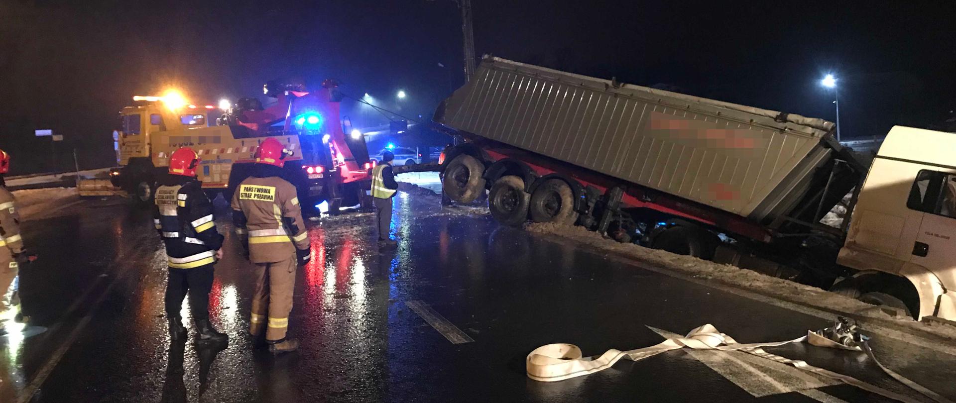 Zdjęcie zrobione w porze nocnej. Holownik pomocy drogowej wyciąga uszkodzony pojazd z rowu. Dwóch strażaków nadzoruje pracę holownika. Po prawej stronie rozwinięta linia gaśnicza.