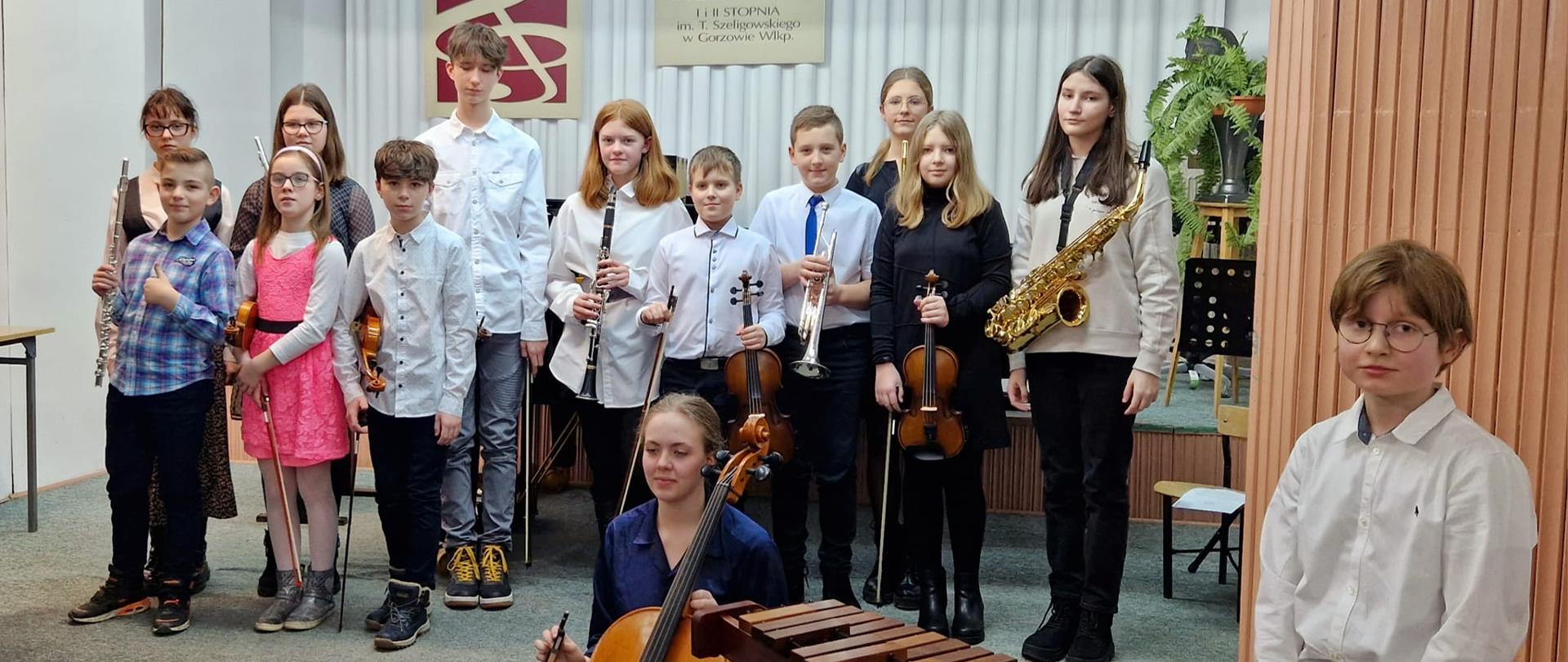 Klasa I cyklu czteroletniego po pierwszym koncercie. Po prawej stronie chłopiec z ksylofonem, obok dziewczynka z wiolonczelą. Na scenie stoją od prawej saksofonistka, skrzypaczka, trębacz, skrzypek, klarnecistka, dwoje skrzypków, flecista. W drugim rzędzie czworo dzieci bez instrumentów.