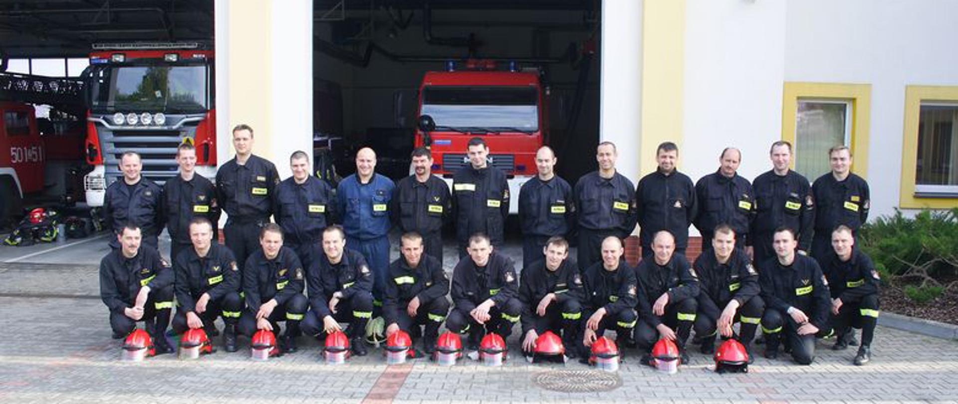 Zdjęcie przedstawia strażaków przed strażnicą z otwartymi bramami w których widać samochody pożarnicze