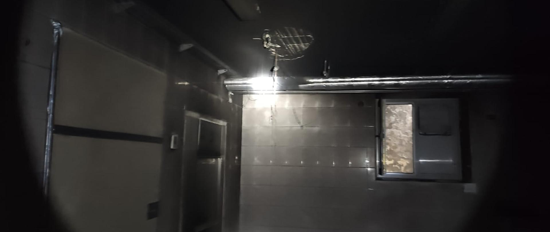 Pomieszczenie piwniczne w którym doszło do zapalenia się lampy sufitowej. Oświetlenie pochodzi z latarki ręcznej. światło skierowane w miejsce spalonej lampy. Całe pomieszczenie odymione.