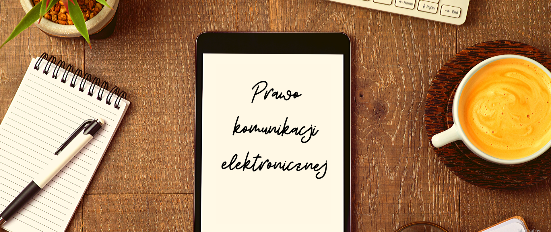 Blat stołu a na nim notes, tablet z napisem "Prawo komunikacji elektronicznej" oraz filiżanka z kawą.