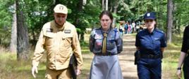 Na zdjęciu widać cztery osoby idące leśną ścieżką w kierunku aparatu. Po lewej stronie znajduje się mężczyzna, strażak w beżowym mundurze, trzymający w ręce czarną teczkę; jego mundur ozdobiony jest naszywkami, a na czapce widnieje godło Polski. Obok niego idzie młoda kobieta w mundurze harcerskim – ma na sobie szarą sukienkę z wieloma przypiętymi odznakami oraz chustę z zielono-czerwonymi paskami. Po jej prawej stronie idzie policjantka w granatowym mundurze z napisem „POLICJA” oraz czapką z orłem. Ostatnia osoba, częściowo widoczna po prawej stronie kadru, również uczestniczy w tej grupie. W tle, wśród drzew, widać namioty i osoby zgromadzone na leśnym obozowisku. Cała scena odbywa się w lesie, w słoneczny dzień, prawdopodobnie podczas inspekcji, wizyty służb na harcerskim obozie.