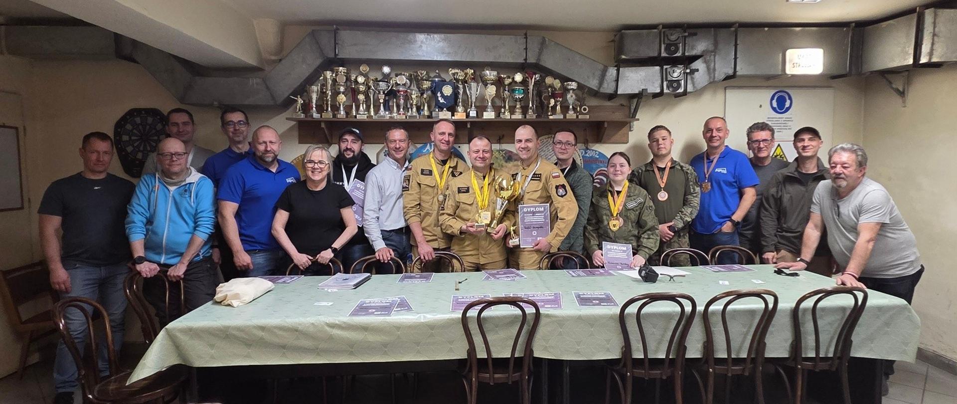 To zdjęcie przedstawia grupę uczestników turnieju strzeleckiego, którzy pozują do wspólnej fotografii wewnątrz strzelnicy. Na pierwszym planie widać długi stół nakryty zielonym obrusem, na którym leżą dyplomy i akcesoria strzeleckie, a w dolnym lewym rogu w kadr wchodzi pies o jasnej sierści. W tle, na półce pod sufitem, dumnie prezentuje się rząd pucharów, co podkreśla sportowy charakter spotkania i tradycję rozgrywanych tu zawodów.
W centrum grupy stoją triumfatorzy turnieju, ubrani w piaskowe mundury, trzymający złote trofea oraz dyplomy. Pozostali uczestnicy są ubrani w stroje sportowe, taktyczne lub codzienne, a wielu z nich ma na szyjach zawieszone medale. Całe pomieszczenie ma surowy, funkcjonalny klimat – widoczne są przewody wentylacyjne, tablice informacyjne dotyczące bezpieczeństwa oraz tarcza do darta, co sugeruje, że jest to miejsce aktywnego wypoczynku i integracji środowiska strzeleckiego.
