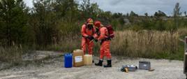 Dwóch ratowników w ubraniach ochronnych bada i zabezpiecza pojemniki z substancjami chemicznymi ustawione na ziemi. Obok znajdują się przyrządy pomiarowe i skrzynki ze sprzętem.
