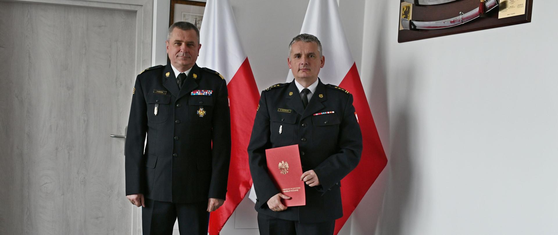Pomorski komendant wojewódzki Państwowej Straży Pożarnej stoi obok strażaka trzymającego czerwoną teczkę. Funkcjonariusze Państwowej Straży Pożarnej ubrani są w mundury wyjściowe z białą koszulą. Za nimi ustawione są dwie flagi Polski na ścianie wisi obraz.