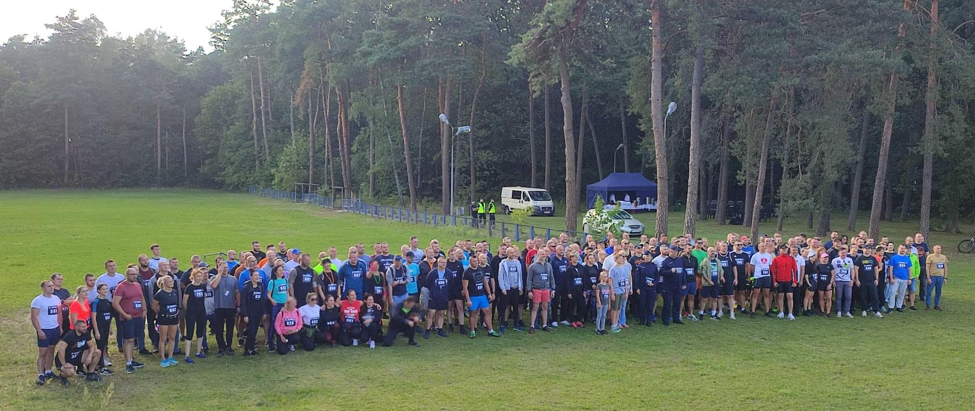Uczestnicy biegu zebrani na leśnym boisku czekający na start, w tle las
