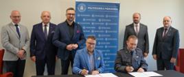 Za przykrytym ciemnoszarym materiałem stołem siedzi dwóch mężczyzn - jeden w niebieskiej marynarce, drugi w ciemnoszarym mundurze, podpisują dokumenty, za nimi stoi pięciu mężczyzn w garniturach, pośrodku grupy minister Kulasek, za nimi pod szarą ścianą niebieski baner z napisem Politechnika Poznańska.