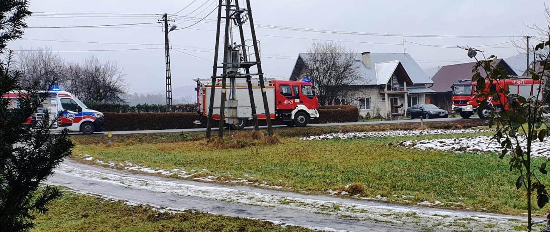 Kolorowe zdjęcie wykonane w pochmurny dzień, przedstawia w oddali drogę, na której stoją pojazdy służb ratowniczych. Od lewej widoczna częściowo, biała karetka pogotowia, z niebiesko-czerwonymi pasami i niebieskim światłem alarmowym na dachu pojazdu. Pośrodku bok czerwonego pojazdu straży pożarnej, stojącego z włączonymi niebieskimi światłami błyskowymi. Po prawej stronie, częściowo przysłonięty krzewami, stojący bokiem, drugi czerwony pojazd straży pożarnej z włączonymi niebieskimi światłami błyskowymi. W tle widoczny jasny budynek mieszkalny a przy nim stojący szary samochód osobowy. Z przodu na zdjęciu droga dojazdowa, szaro-zielona trawa i miejscowo leżący niewielki śnieg.