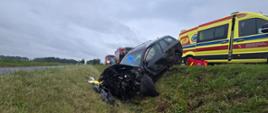 Czarny samochód w rowie, mocno uszkodzony przód, po prawej karetka na drodze