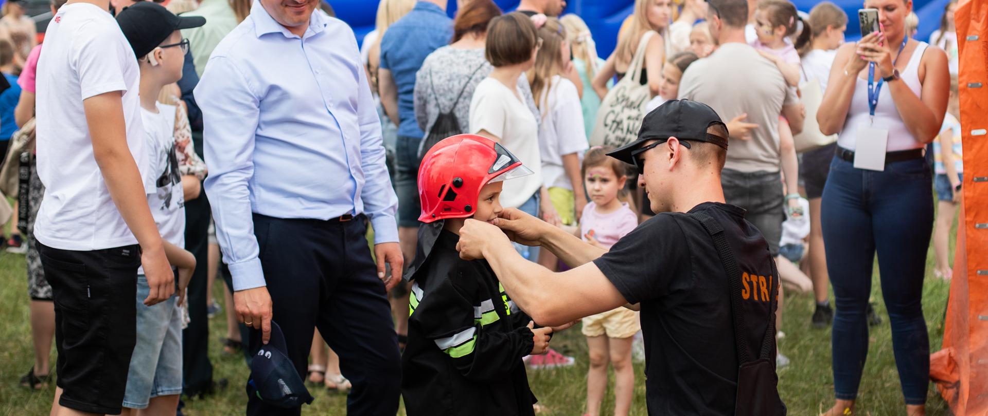 Strażak ubiera małego chłopca w mundurek strażacki. Chłopiec ma na głowie czerwony hełm. Wokół przyglądają się temu ludzie, a w tle dmuchana ślizgawka.