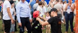 Strażak ubiera małego chłopca w mundurek strażacki. Chłopiec ma na głowie czerwony hełm. Wokół przyglądają się temu ludzie...