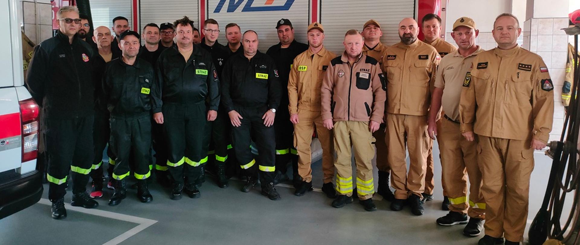 W garażu, na tle samochodu ratowniczo-gaśniczego w kolorze czerwonym stoi grupa mężczyzn pozując do zdjęcia. Część z nich ubrana jest w czarne ubrania koszarowe, część w ubrania w kolorze piaskowym.