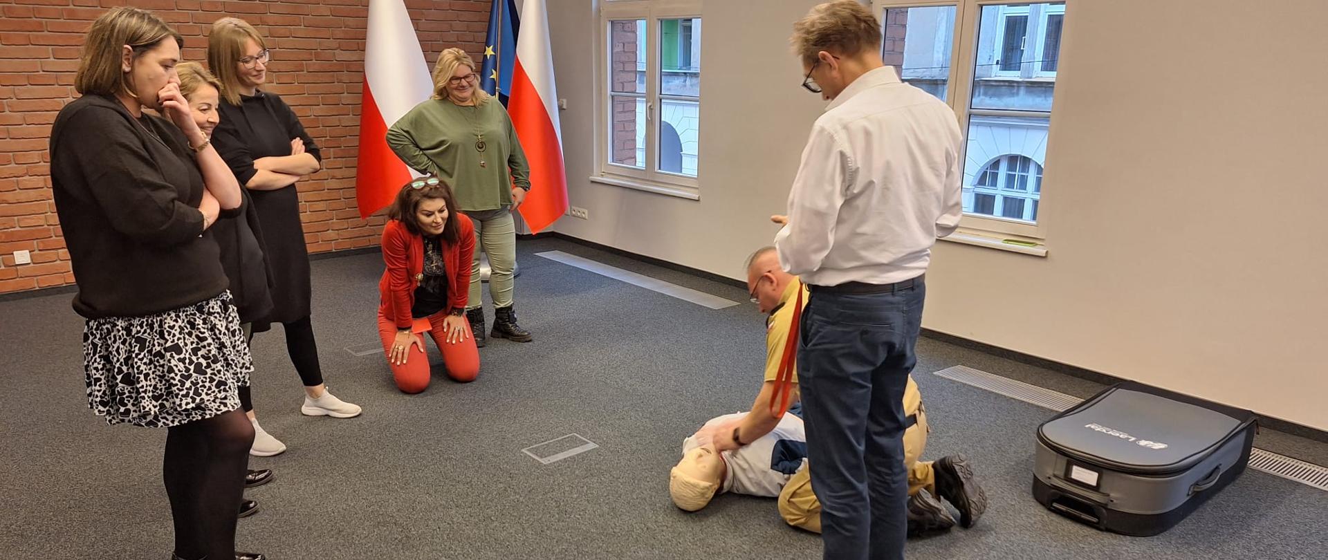 w dużej sali na ziemi strażak klęczy przy manekinie, cztery kobiety i mężczyzna przyglądają się strażakowi