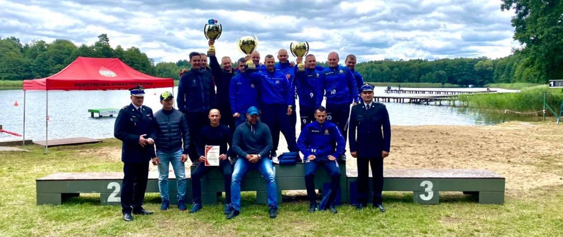 Zwycięzcy drużynowych zmagań podczas XXVI Mistrzostw Polski Strażaków Płetwonurków, wraz z nimi Zachodniopomorski Komendant Wojewódzki st. bryg. Jarosław Tomczyk (po lewej w mundurze) oraz naczelnik Wojewódzkiego Ośrodka Szkolenia bryg. Tomasz Dębowski (po prawej w mundurze). W tle widoczne jezioro Pile