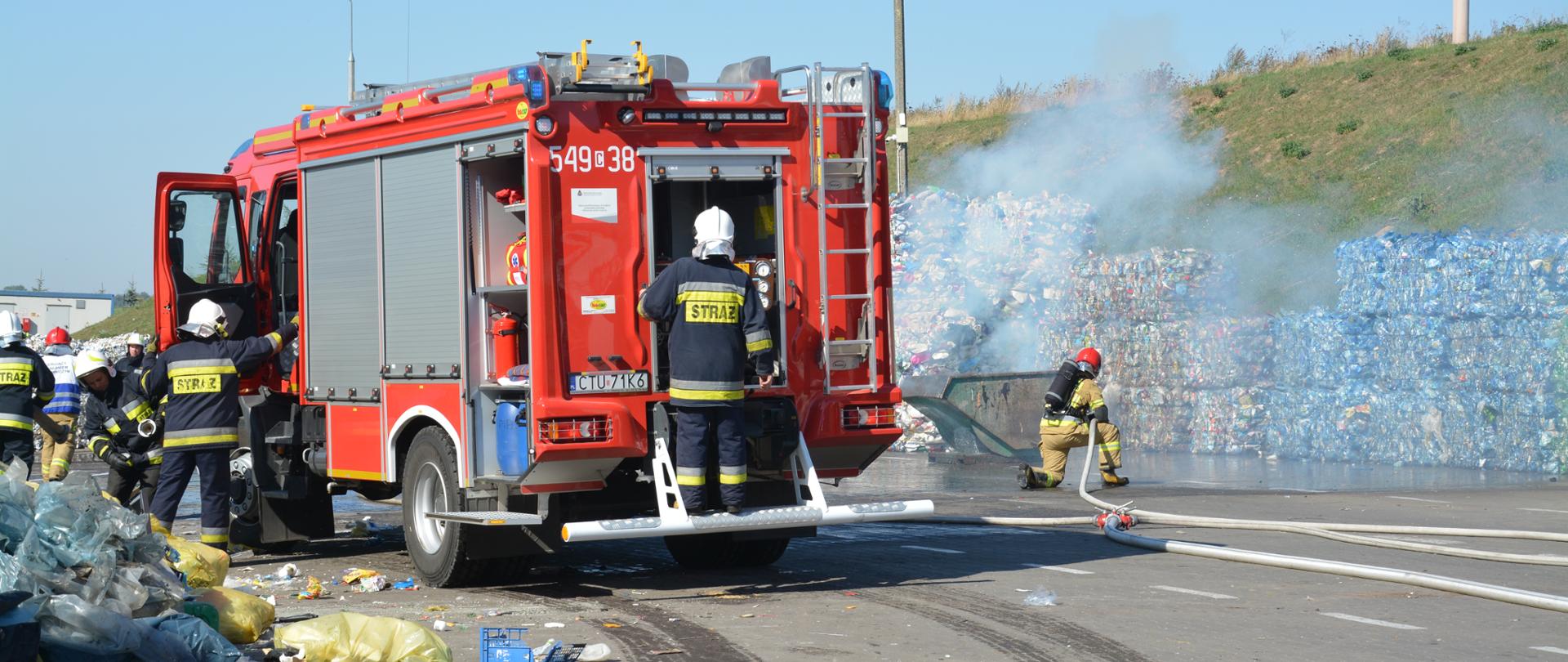 Na zdjęciu widać strażaków w ubraniach specjalnych prowadzących rozwinięcie bojowe. Na pierwszym planie samochód ratowniczo-gaśniczy. W tle widać dym i strażaków gaszących pożar odpadów komunalnych. 