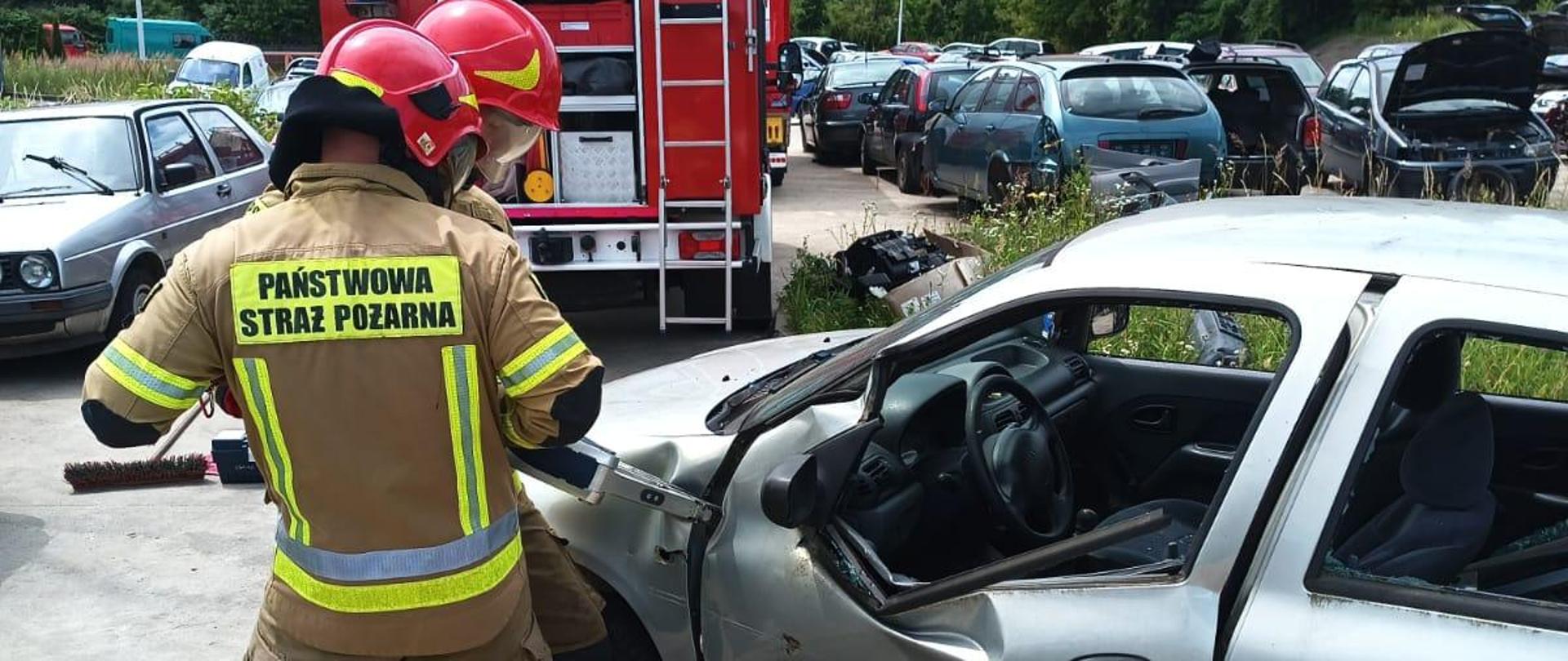 Dwóch strażaków robi dostęp do wnętrza srebrnego wraku pojazdu poprzez usunięcie drzwi.