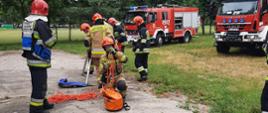 Zdjęcie przedstawia sześciu strażaków sprawiających sprzęt do ewakuacji ze studni i wykopów. W tle dwa samochody strażackie.