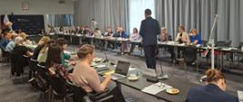 Zdjęcie przedstawia jasną salę. Po lewej stronie w tle widać ściankę Ministerstwa Rozwoju i Technologii. W sali siedzą uczestnicy spotkania przy stołach, które są ustawione w podkowę. Na stołach stoją mikrofony oraz standy z nazwami urzędów.