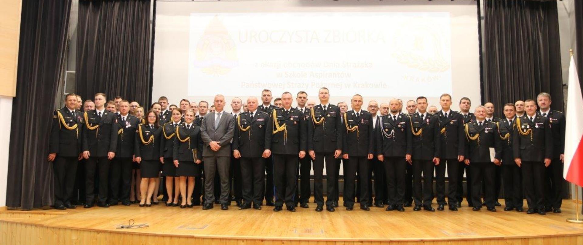 Zdjęcie grupowe uczestników zbiórki okazji Dnia Strażaka w Szkole Aspirantów Państwowej Straży Pożarnej w Krakowie. Osoby stoją na scenie w auli, są wśród nich pracownicy i funkcjonariusze szkoły oraz zastępca komendanta głównego Państwowej Straży Pożarnej,