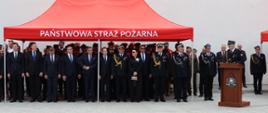 Goście uroczystości stoją pod czerwonym namiotem z napisem „PAŃSTWOWA STRAŻ POŻARNA”. 