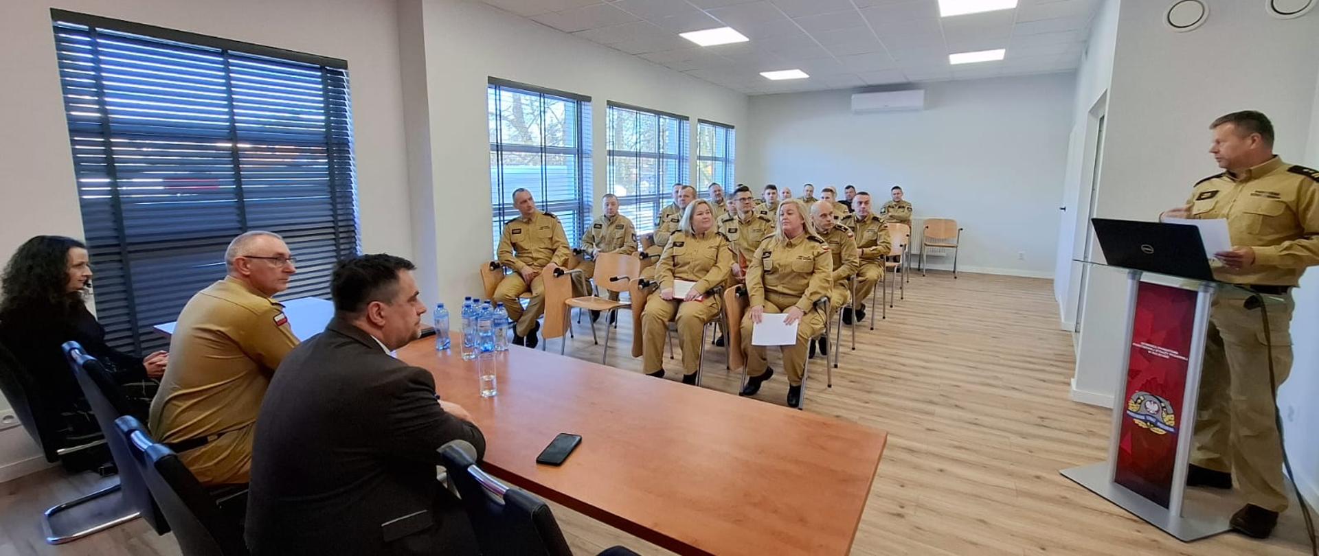 Komendant Powiatowy wita gości, po lewej przy stole Komendant Wojewódzki i Starosta, w głębi funkcjonariusze na krzesłach