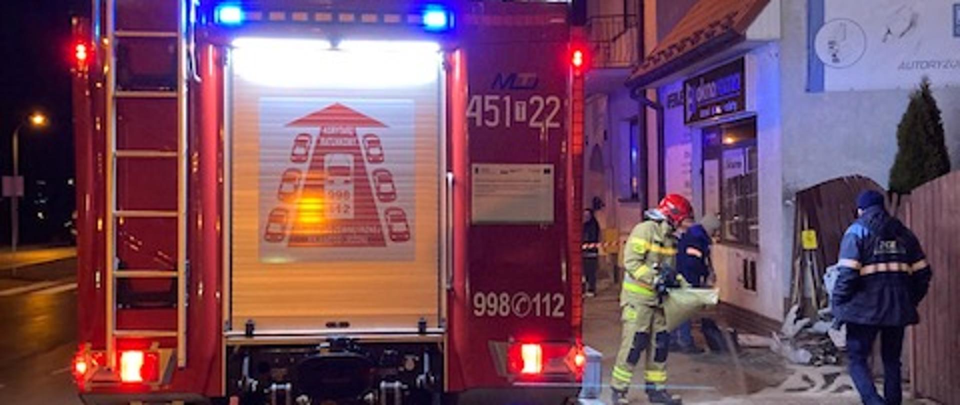 Na zdjęciu w porze nocnej widać na pierwszym planie oświetlony tył samochodu ratowniczo-gaśniczego. Po prawej stronie samochodu idzie jeden w pracowników zakładu energetycznego oraz strażak, który usuwa wyciekłe płyny eksploatacyjne z pojazdu, który uderzył w skrzynkę elektryczną.