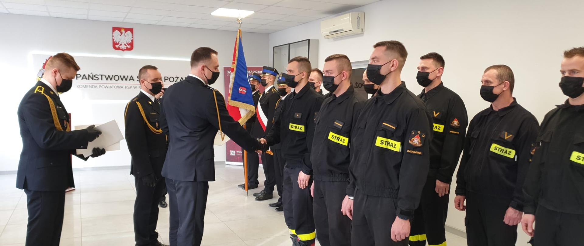 Świetlica komendy. Komendant powiatowy gratuluje jednemu strażakowi stojącemu w szeregu. W tle sztandar i ściana z logo PSP. U góry godło Polski.