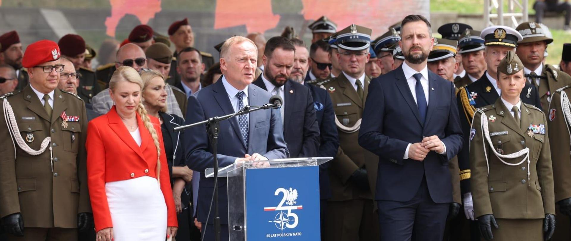 Wiceminister Czesław Mroczek stoi przy mównicy. Po lewej kobieta w czerwonej marynarce, po prawej wicepremier Kosiniak-Kamysz. W tle osoby ubrane w mundury żołnierskie 