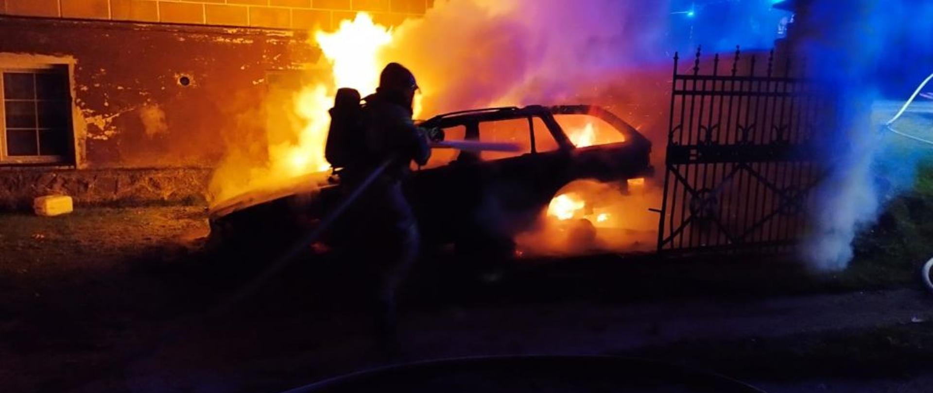 Zdjęcie wykonane w nocy. Centralny punkt zdjęcia stanowi pomarańczowy kolor, ogień obejmujący auto. Zdjęcie ciemne. Widać tylko zarys palącego się ognia i gaszącego je strażaka.
