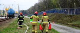 Wagon stoi na bocznicy kolejowej w niedalekiej odległości rozwinięte są węże pożarnicze. Na pierwszym plamie widzimy 3 strażaków niosących pachołki oraz węża pożarniczego. W oddali obok wagonu widoczni są 2 strażacy prowadzący działania. 