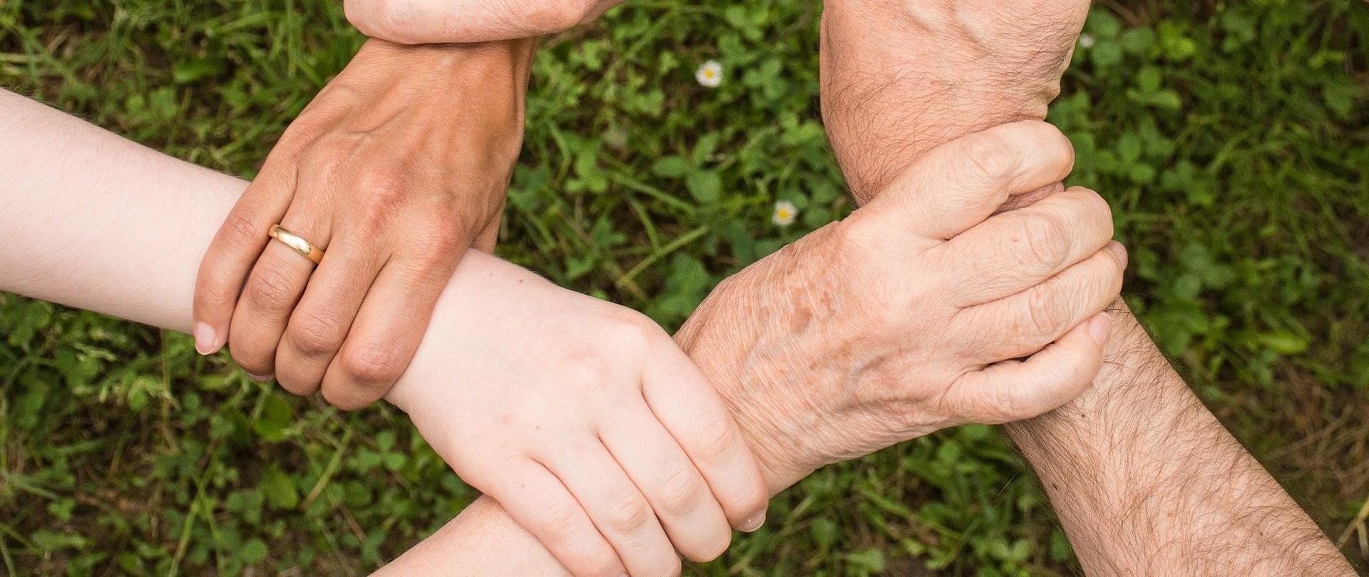 Na zdjęciu trzy pary dłoni, trzymające się w uścisku w kształcie kwadratu. W dalszej części planu trawa. Zdjęcie zrobione z góry. 