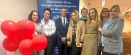 Pani Dyrektor WSSE we Wrocławiu na sali wykładowej wraz z pracownikami. W tle baner Inspekcji Sanitarnej oraz czerwone balony