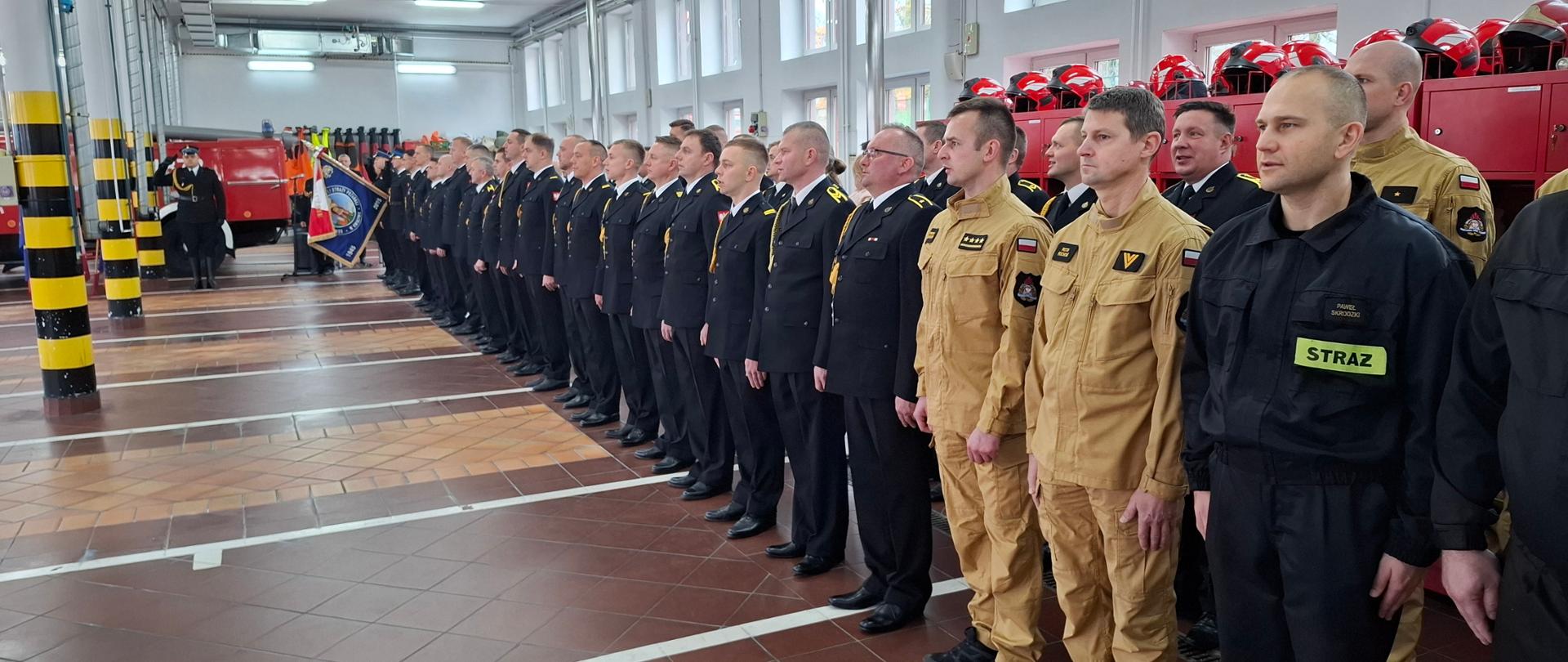 Strażacy stoją w dwuszeregu, na początku szyku poczet sztandarowy
